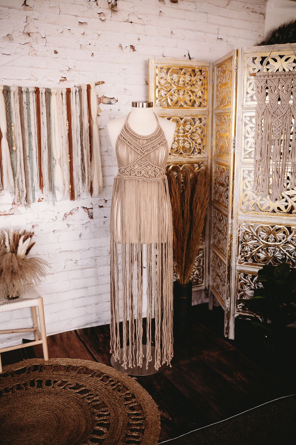 A beige macrame fringe halter dress displayed on a mannequin in a boho-styled studio with woven wall decor.
