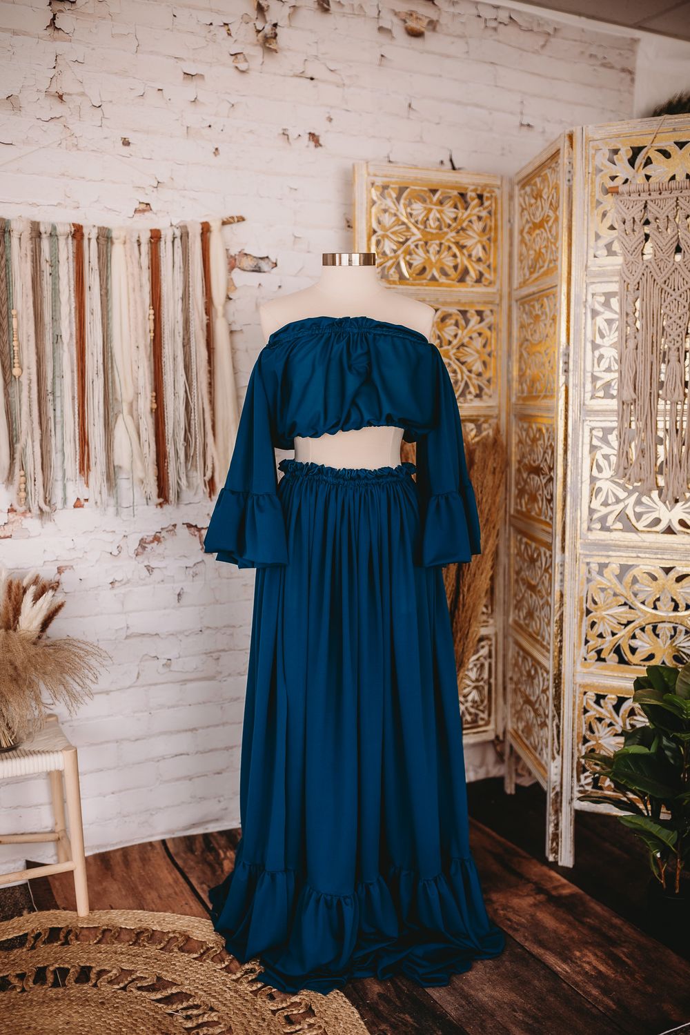 A teal blue off-shoulder crop top and matching ruffled maxi skirt set on a mannequin in a boho studio.
