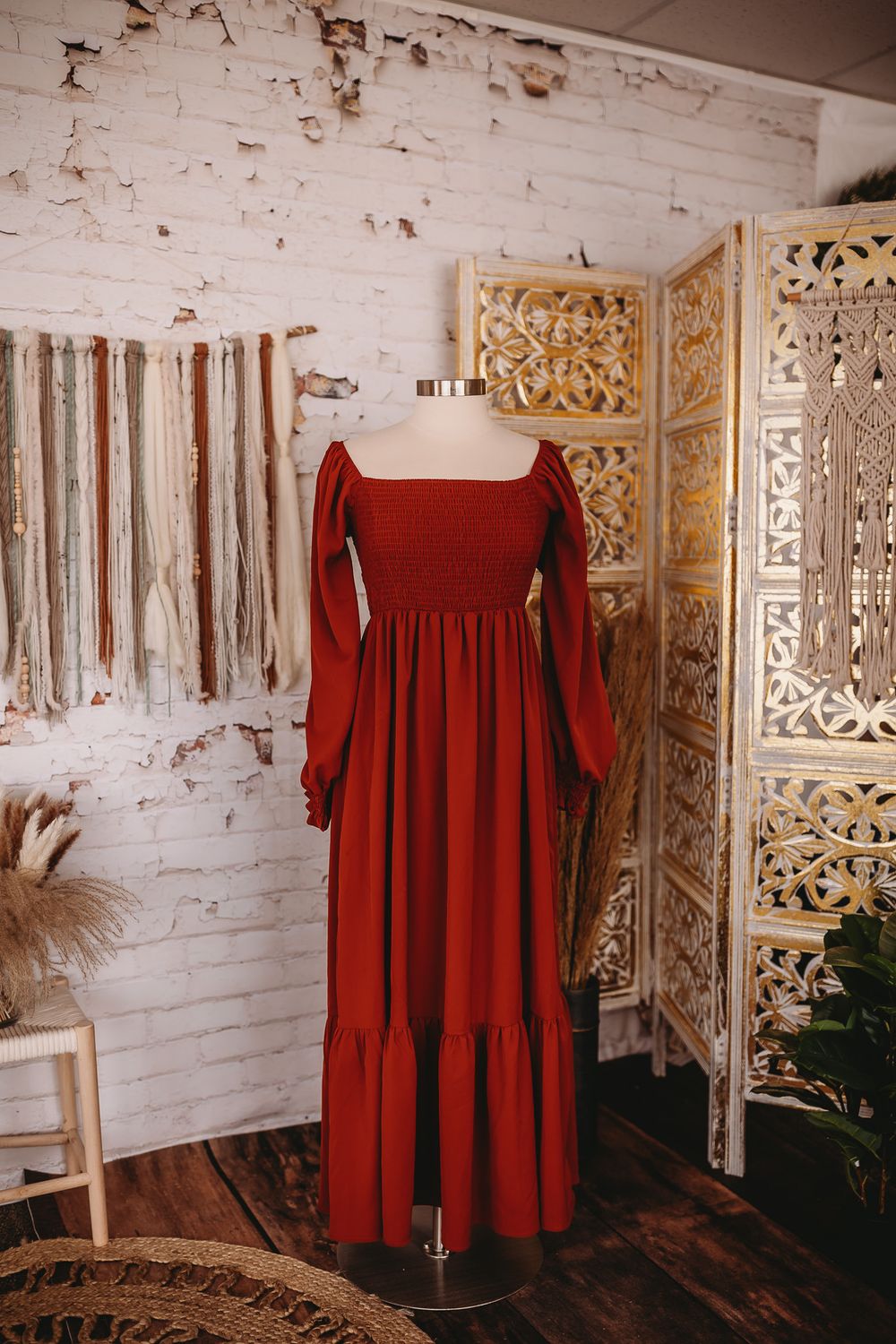 A rust red smocked long-sleeve maxi dress with bell sleeves displayed on a mannequin in a boho studio.