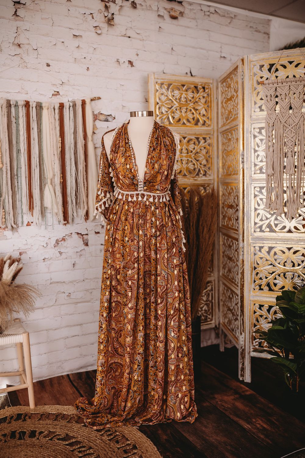A deep brown paisley print halter maxi dress with tassel belt displayed on a mannequin in a boho studio.