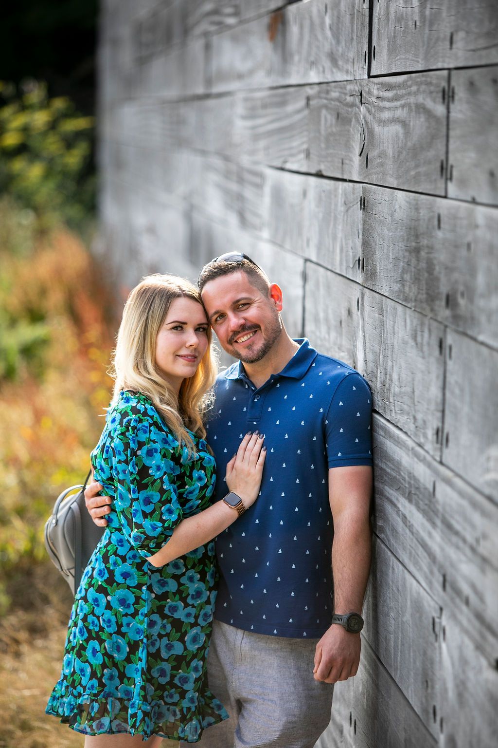 A couple poses together against a gray wall, wearing a blue floral dress and polka dot polo shirt.