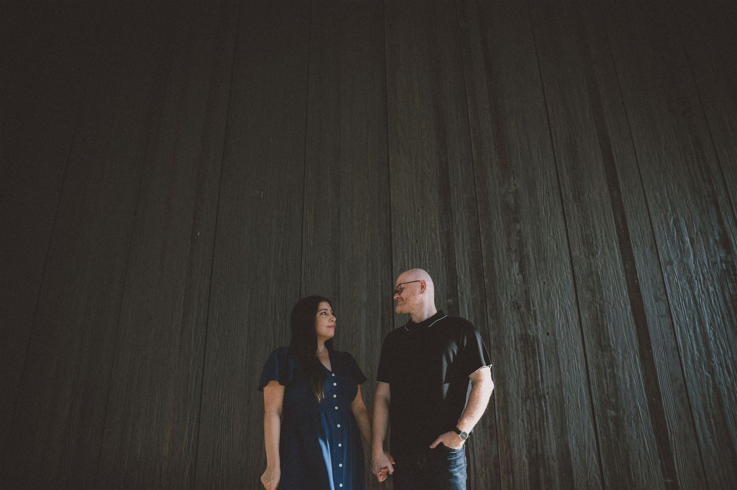 A couple stands against a dark textured wall in contrasting outfits.