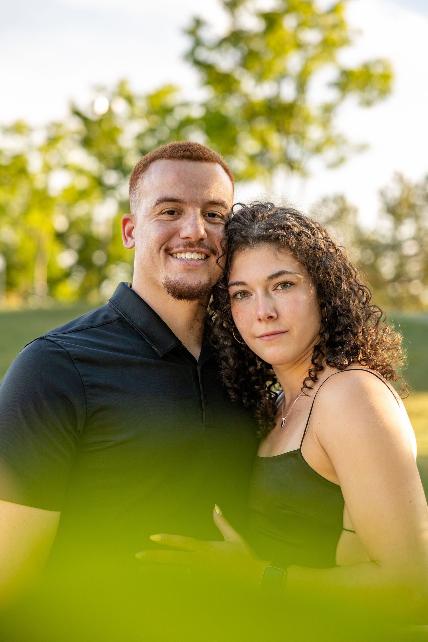 A summer portrait of a couple framed by soft green bokeh leaves in golden sunlight.