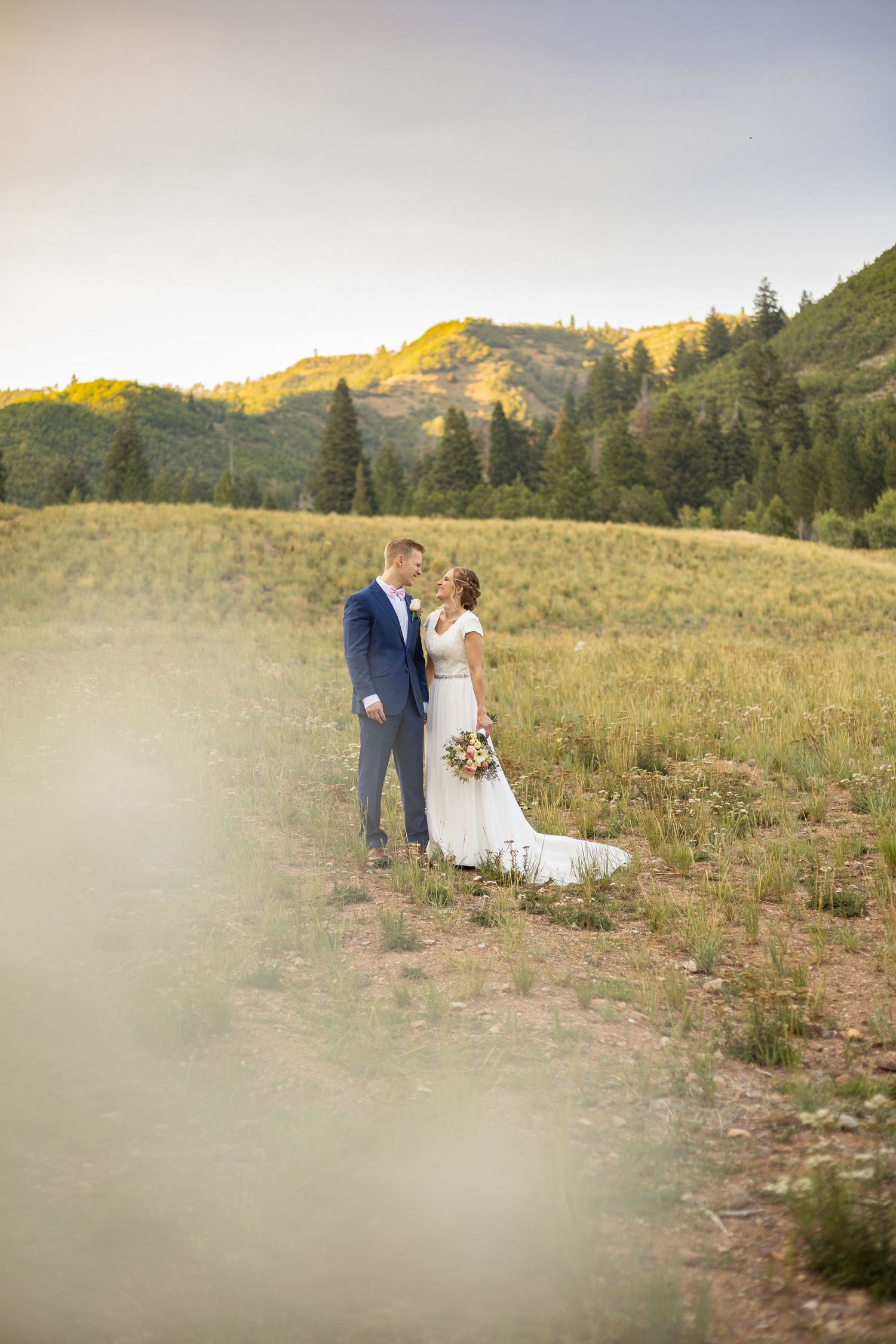 A couple in wedding attire embraces in a mountain meadow at sunset with evergreen trees and golden hills in background.