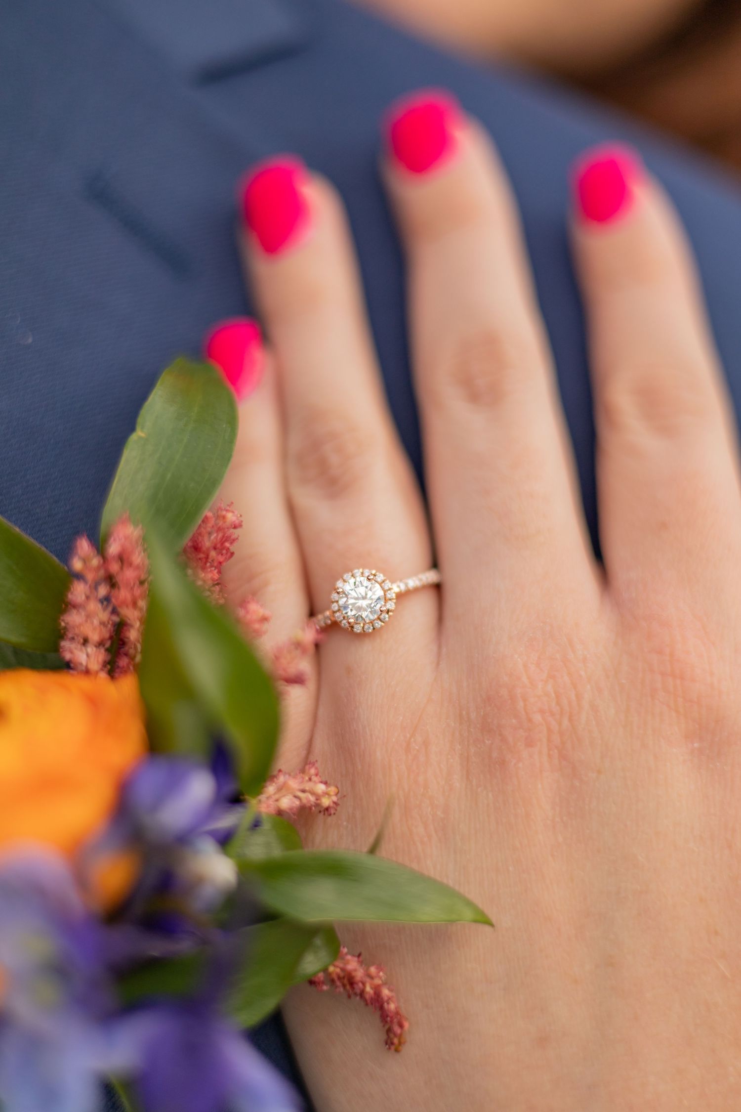 A close-up of an engagement ring on a hand with bright pink nails and colorful flowers.