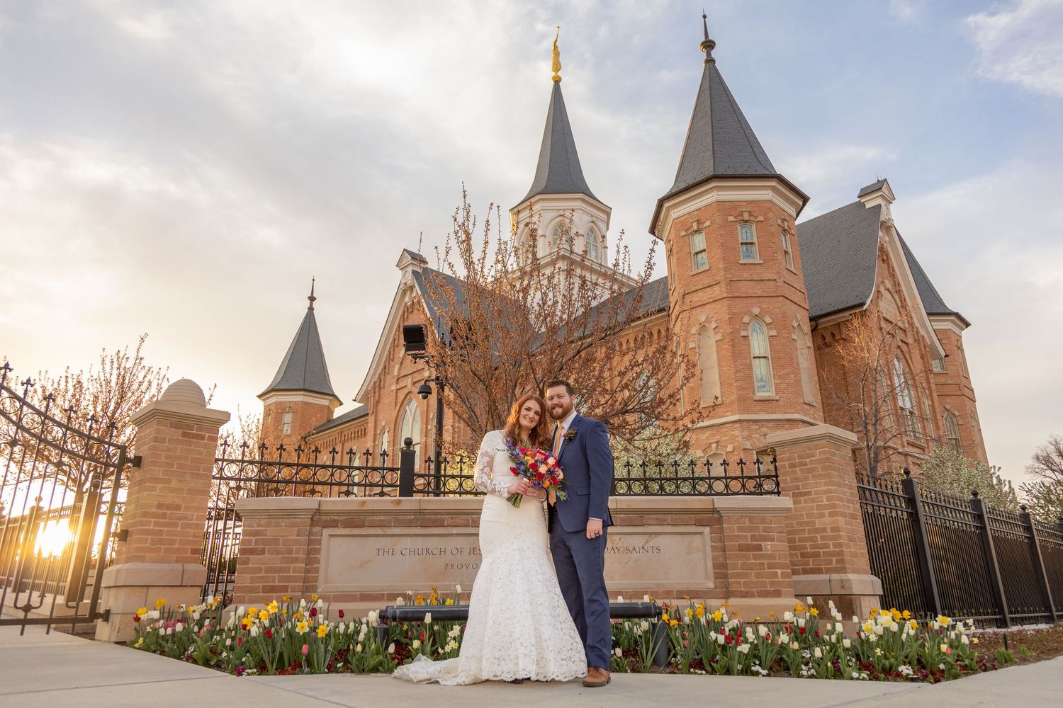 A couple poses in front of a grand castle-like building with spires at sunset surrounded by spring flowers.