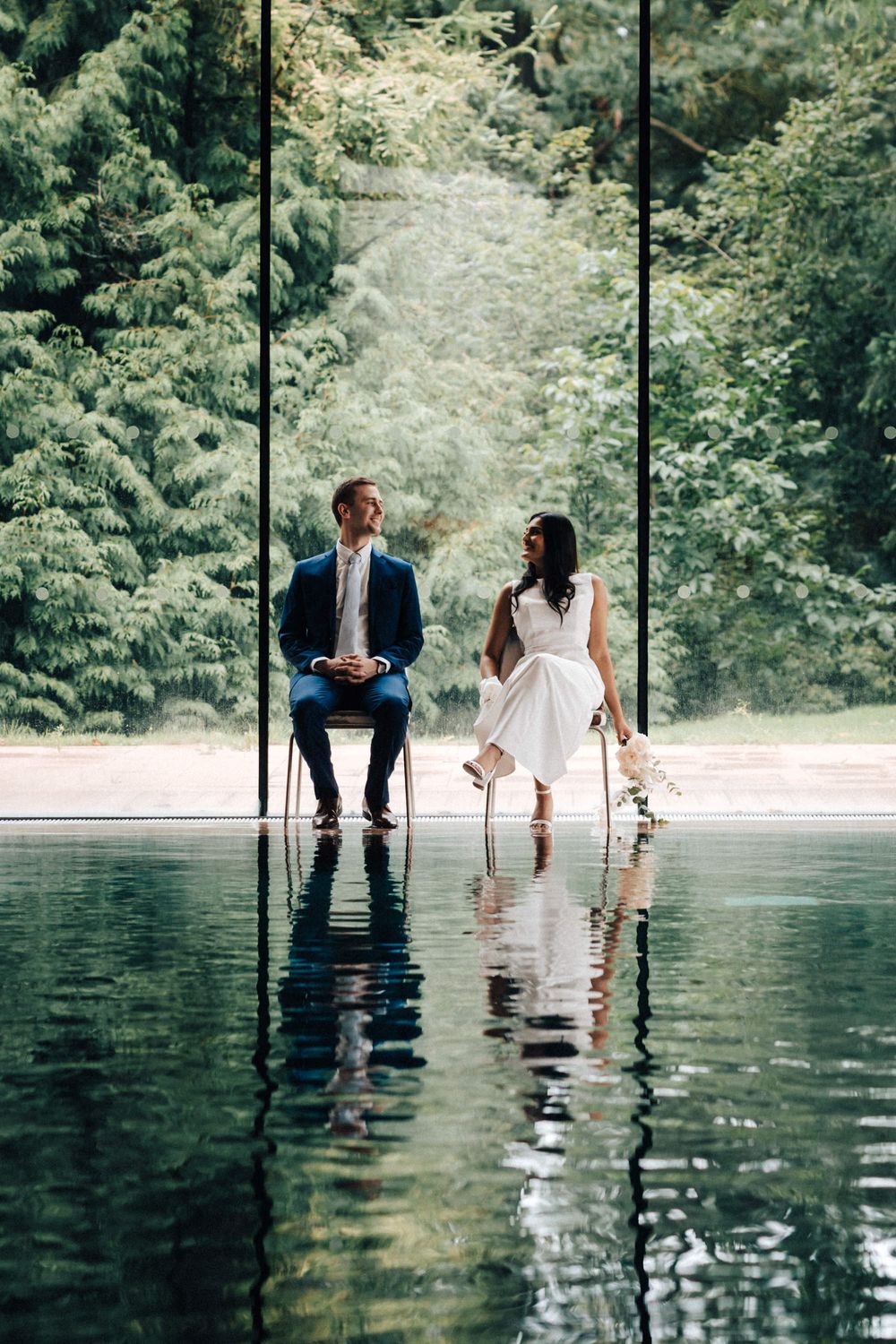 Bride and groom sitting on chairs at the swimming pool looking at each other with reflections