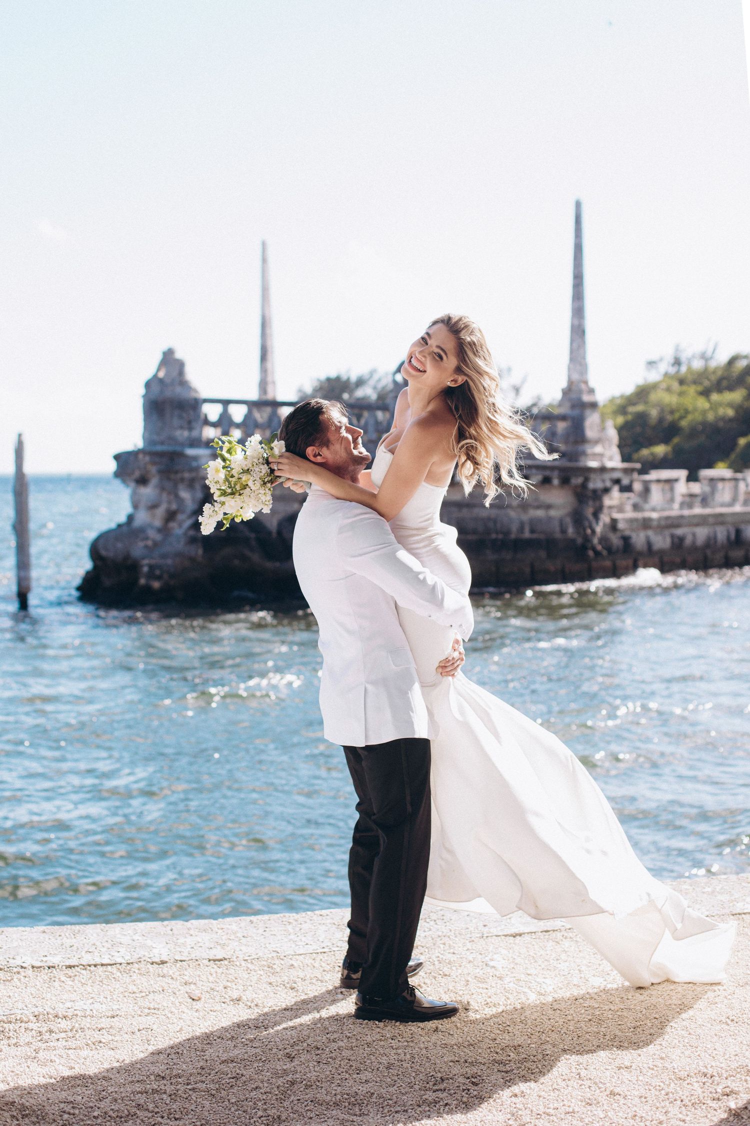 A joyful seaside wedding moment captured near historic stone architecture and sparkling water.