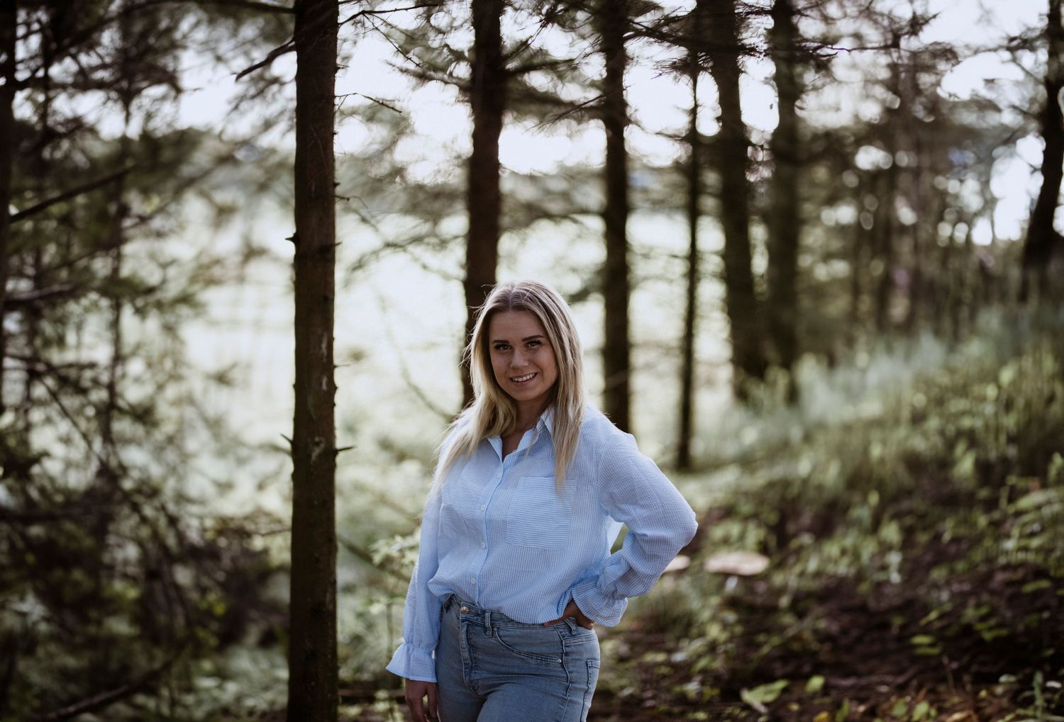 A woman in a blue shirt and jeans poses in a forest setting during golden hour.