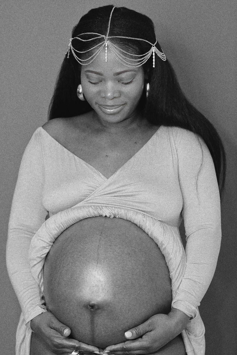 A pregnant woman in white dress wearing a delicate chain headpiece smiles while cradling her baby bump.