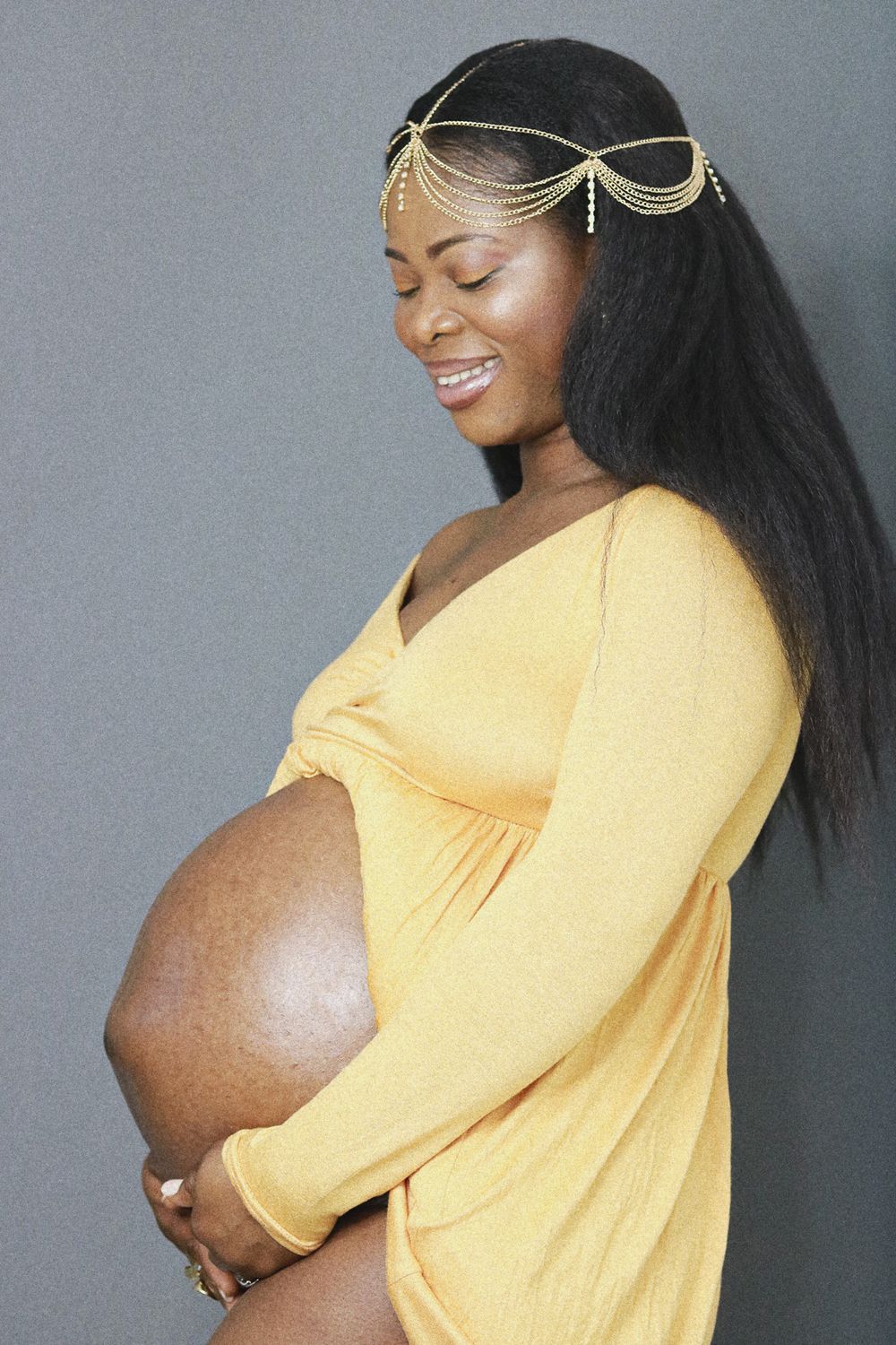 An expectant mother in yellow dress poses with hands on belly while wearing a decorative gold headchain.