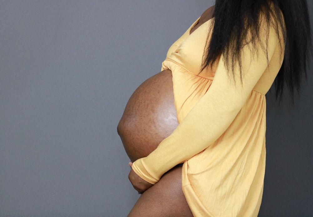 Side view of pregnant belly in yellow dress against grey background.