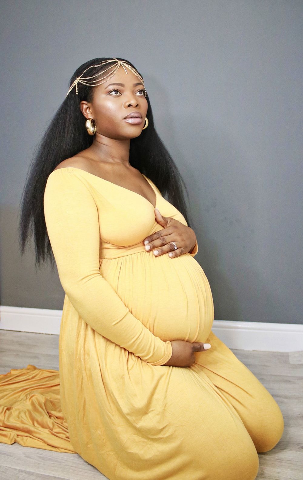 Elegant maternity portrait of woman in yellow dress kneeling on floor with hands on belly.