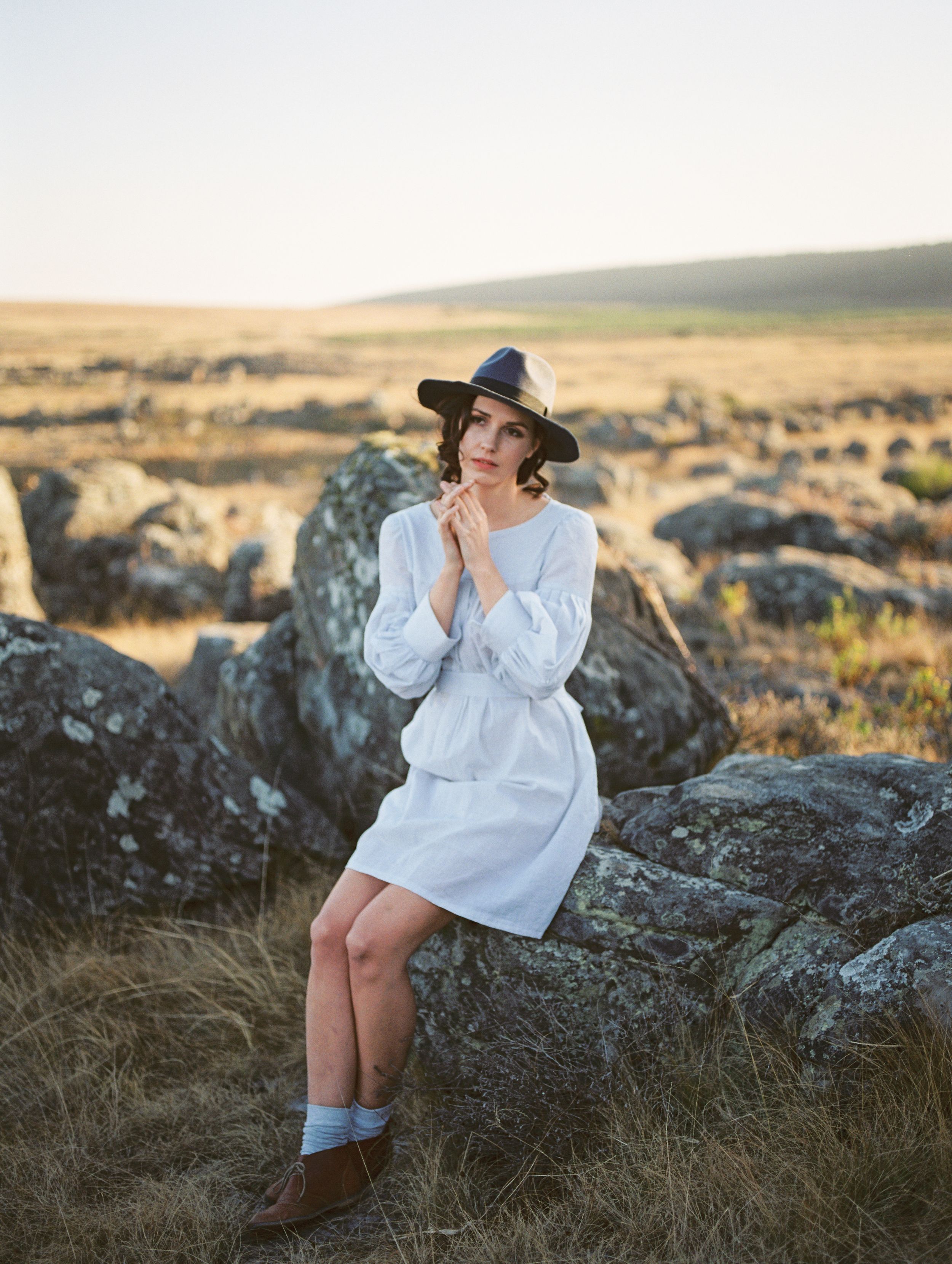 Daintily holder her face a woman poses on rocky outcrops wearing a long sleeve dress and hat.