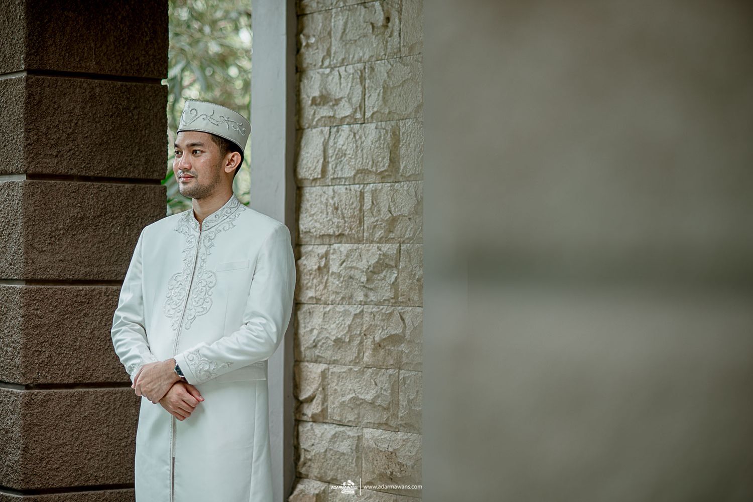 A groom in traditional Islamic dress stands thoughtfully against a stone wall.
