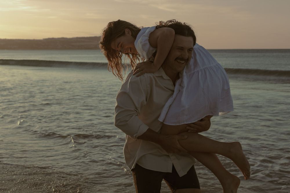 Two people embrace while standing in shallow ocean water during golden hour.