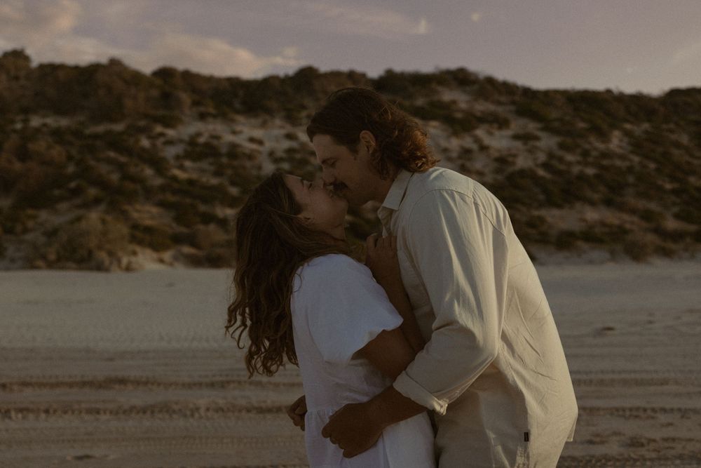 Couple sharing an intimate moment on a beach with rocky cliffs in the background.