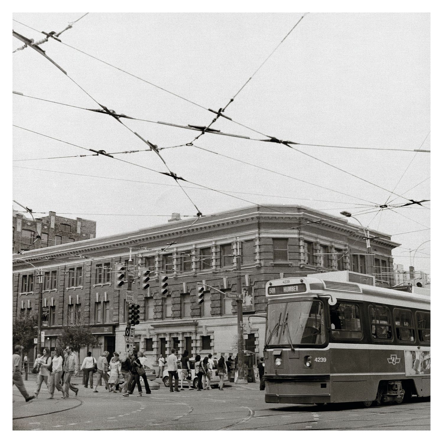 Black and white urban scene with streetcar and pedestrians beneath overhead power lines.