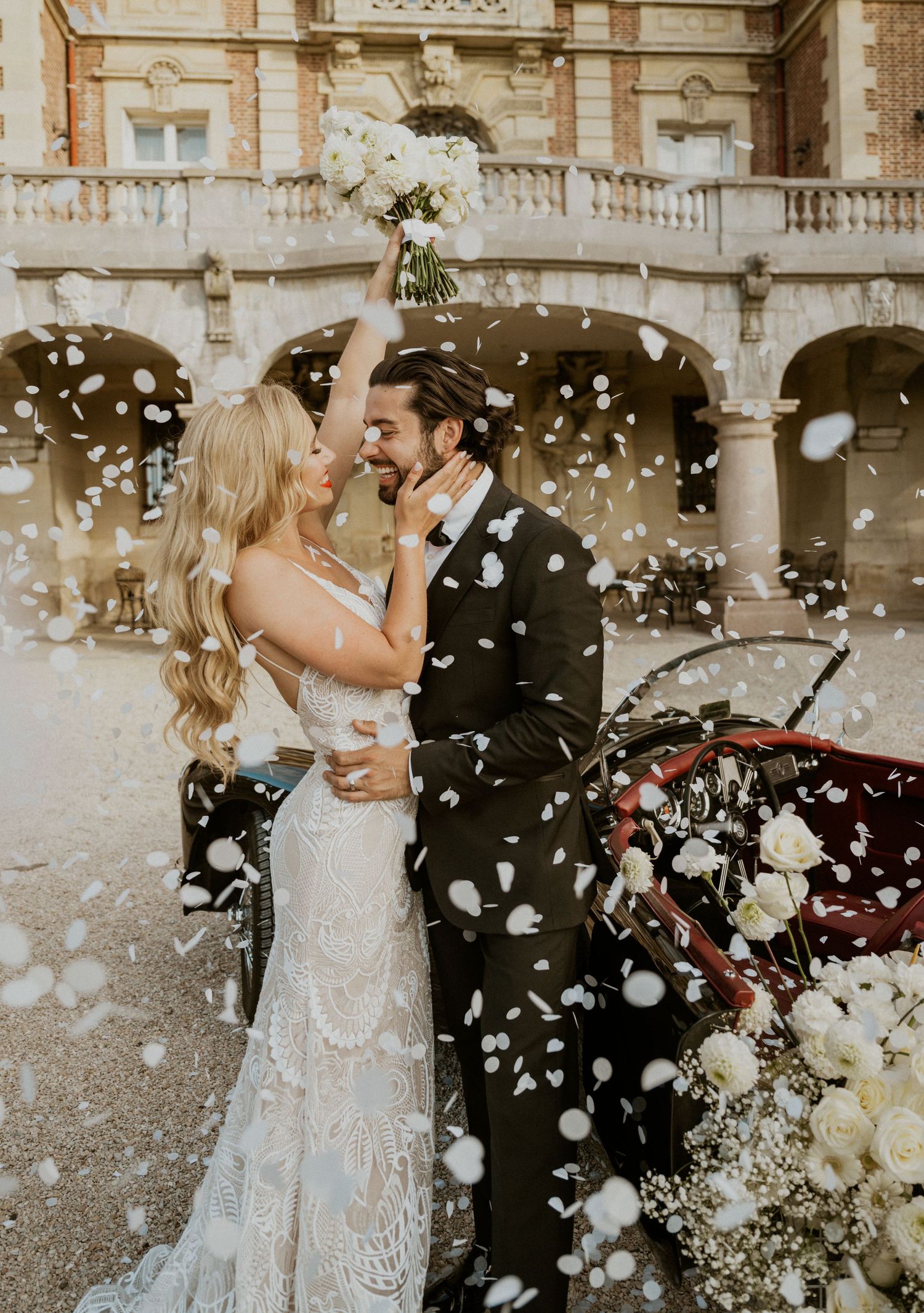 Romantic wedding couple embraces as flower petals fall around them at a grand historic venue.