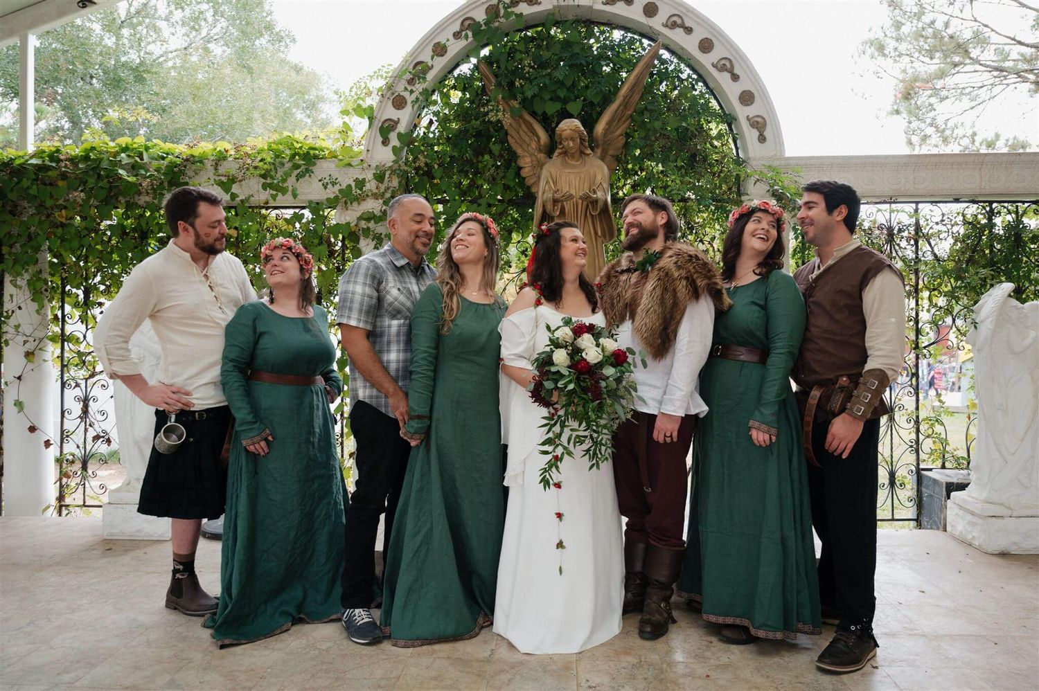 A wedding party in medieval attire and emerald green dresses poses under a decorated arch.