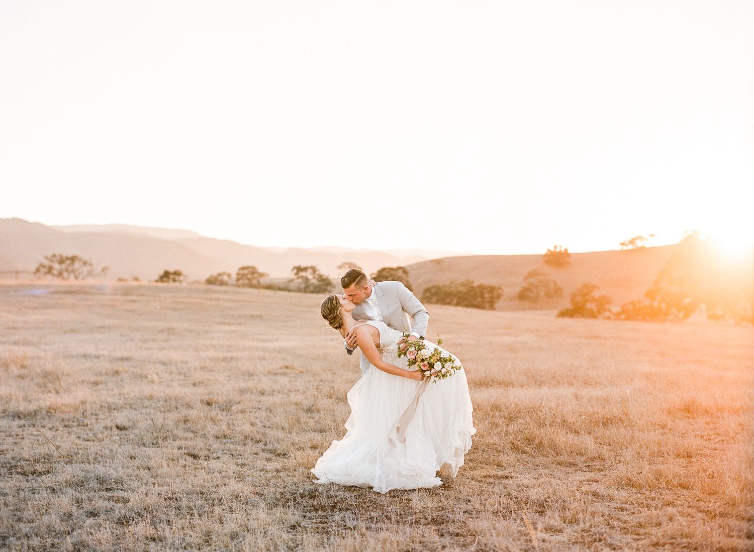 Bride and groom with bouquet kissing in a field at sunset at Kestrel Park wedding