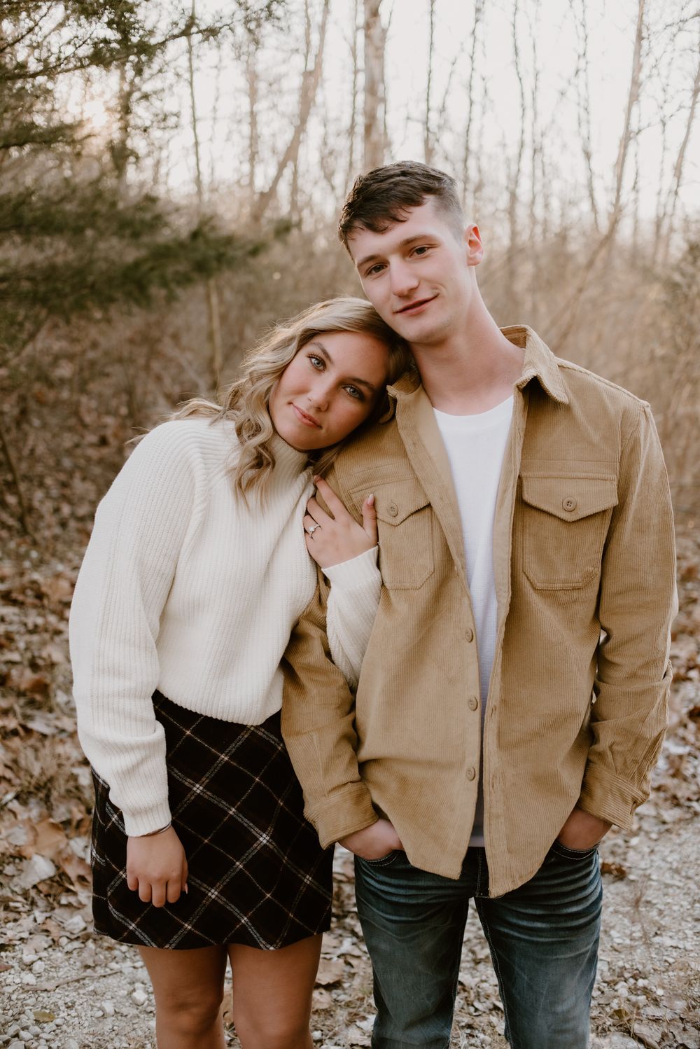 Couple poses together against bare trees in autumn, wearing white sweater and tan jacket.