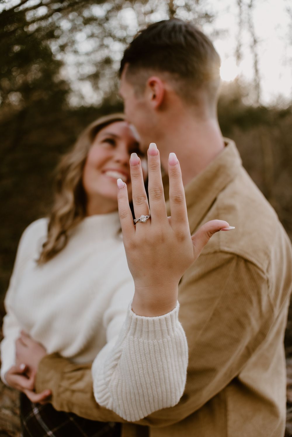 A hand displays an engagement ring while couple embraces in a forest setting.