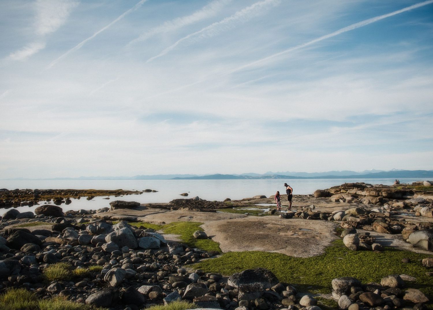 Rocky coastal landscape with distant mountains and blue waters under wispy clouds.