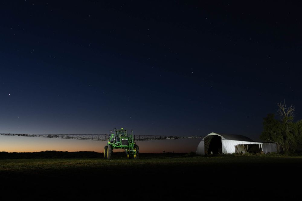 Starlit night sky over silhouetted farm buildings and equipment on the horizon.