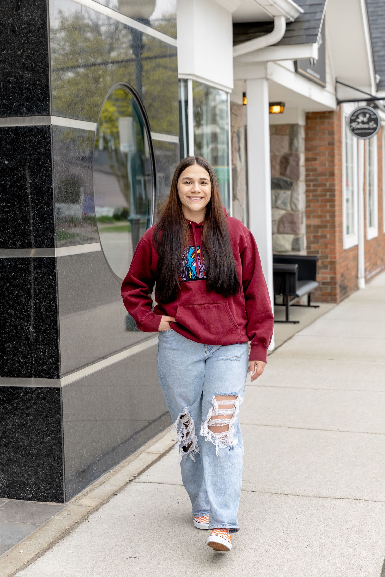 teenager in distressed jeans and a burgundy sweatshirt walking down a sidewalk in Rochester Michigan for senior photos