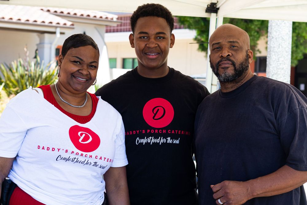 Three people wearing casual t-shirts smile together at an outdoor event.