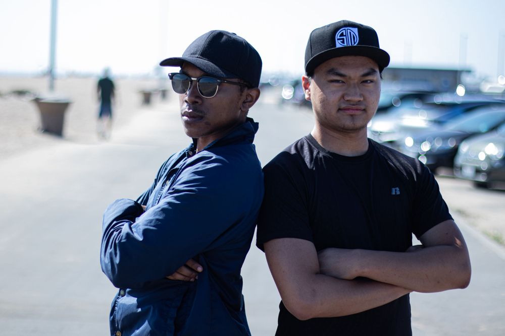 Two friends posing back-to-back outdoors wearing casual attire.