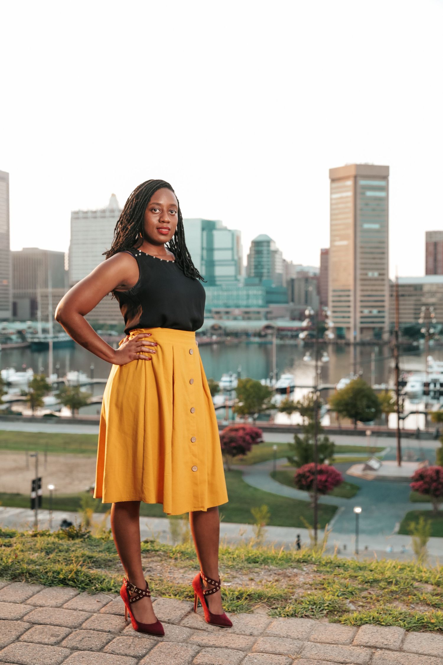 Confident woman in yellow midi skirt and red heels posing with city skyline harbor backdrop.
