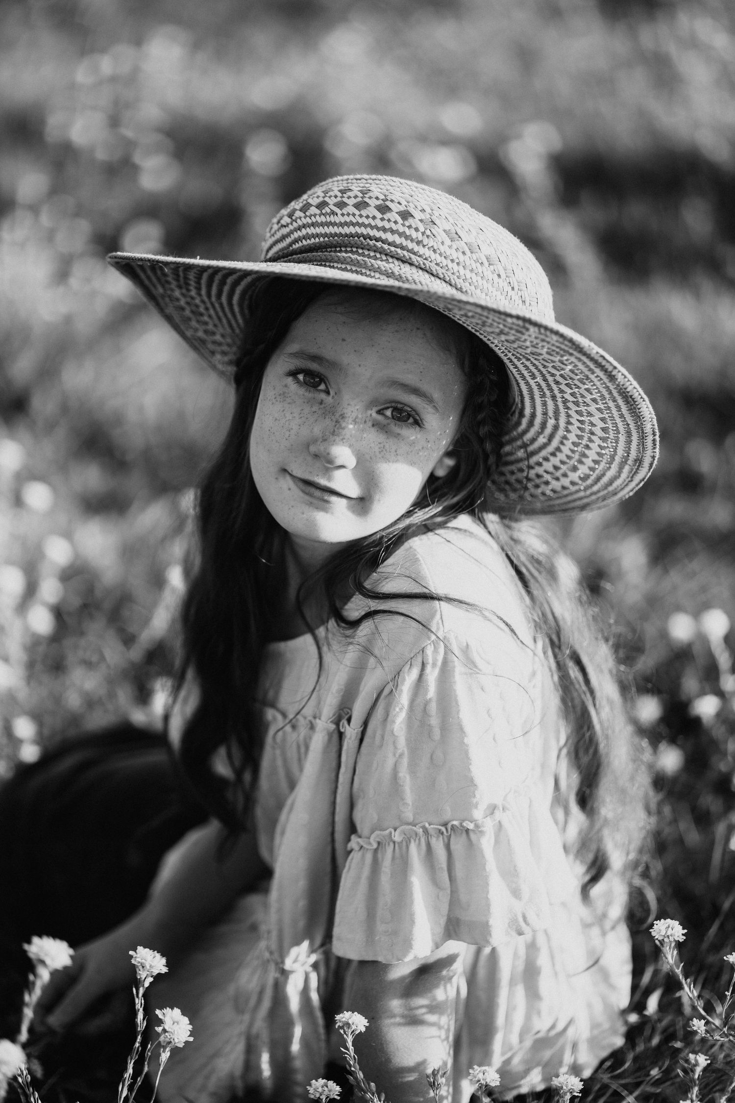 A young girl in a sun hat poses playfully among wildflowers in a black and white portrait series.