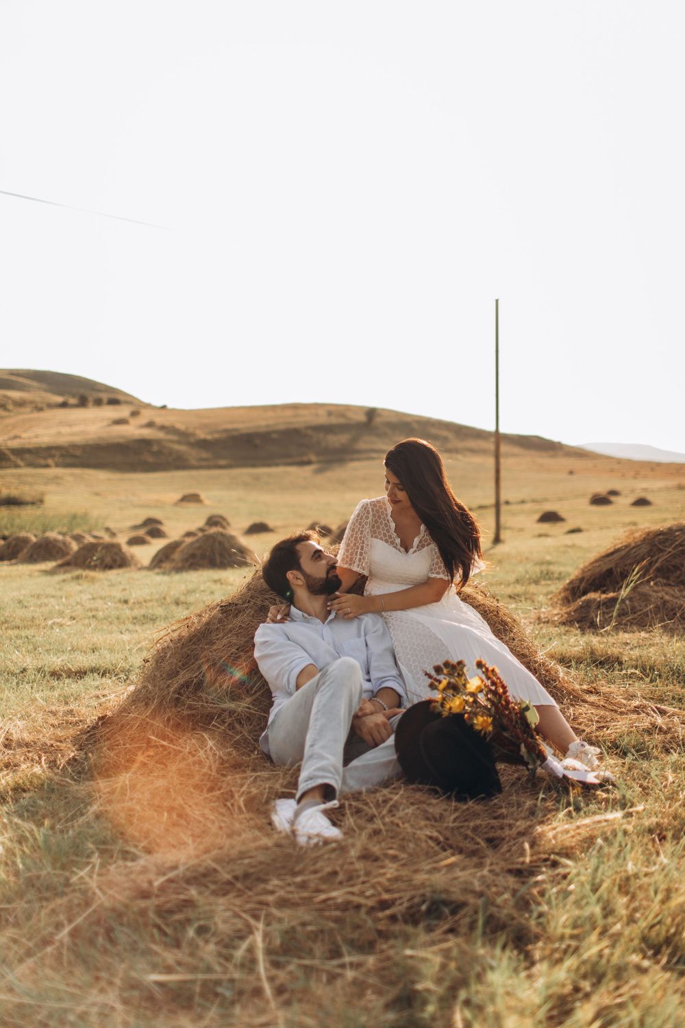 Couple relaxing on hay bale with sunflowers against golden hills at sunset.