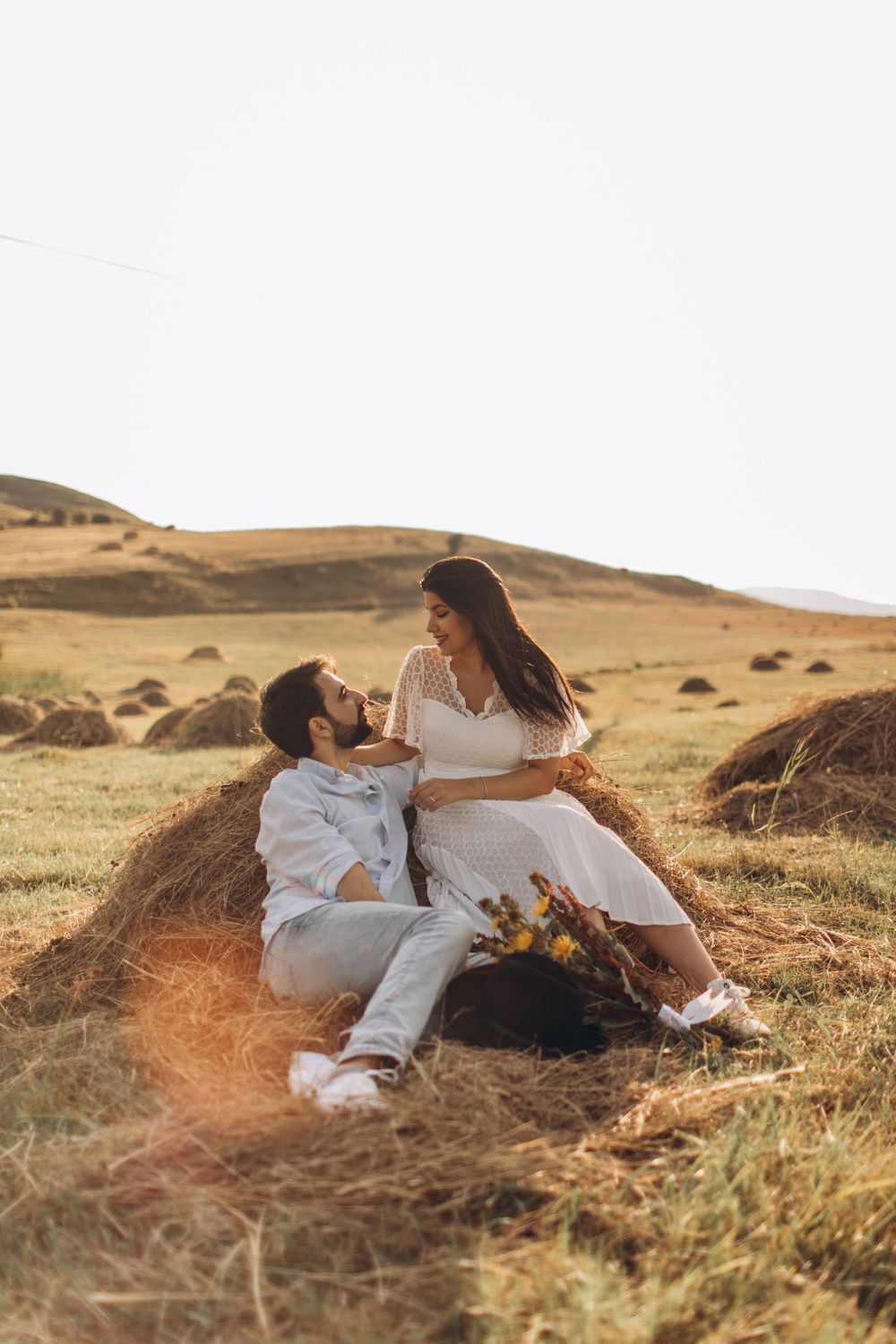 Romantic couple relaxing on hay bales in golden field during sunset photoshoot.