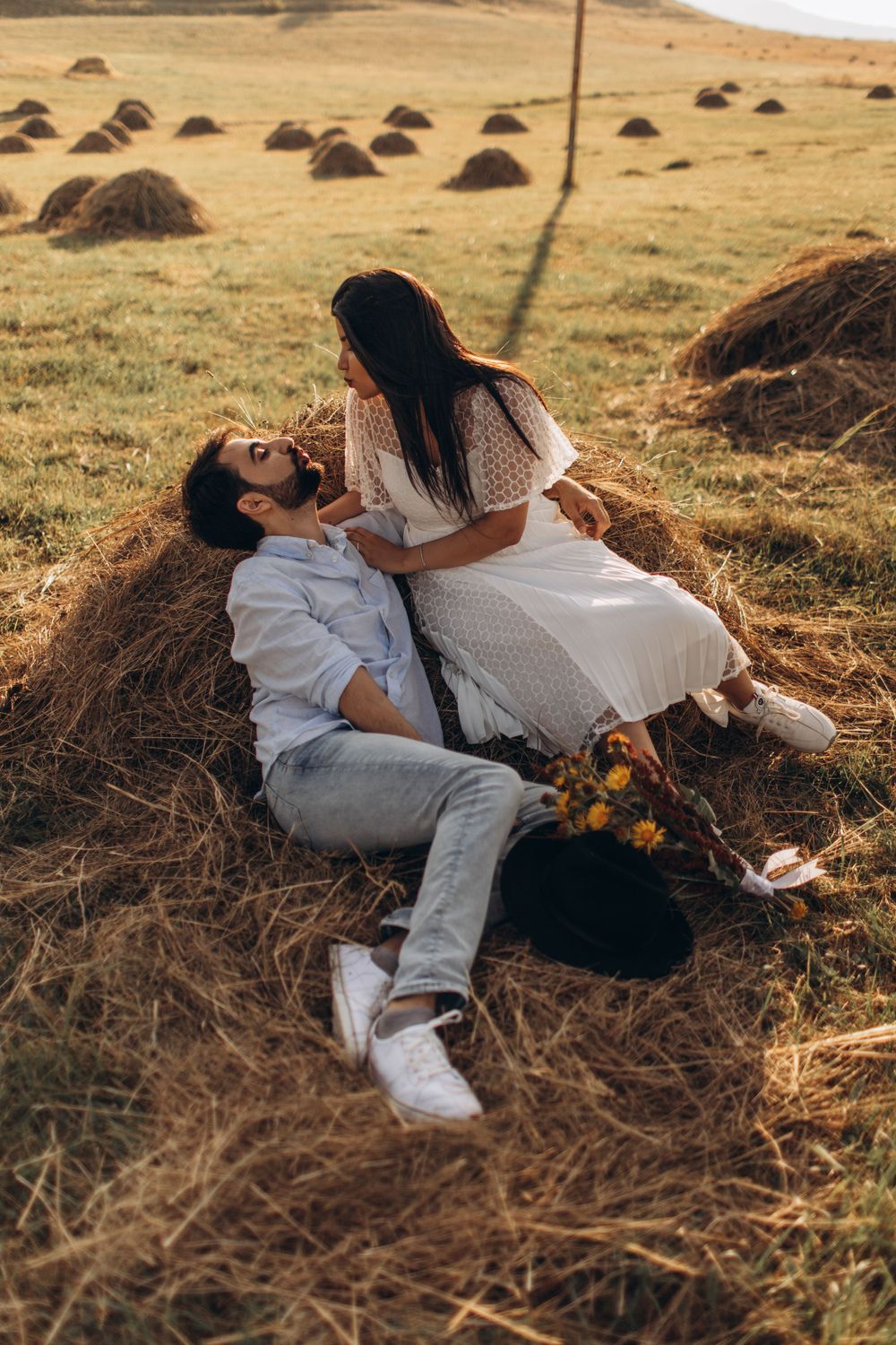 Couple sharing tender moment while resting against hay bale in rustic field setting.