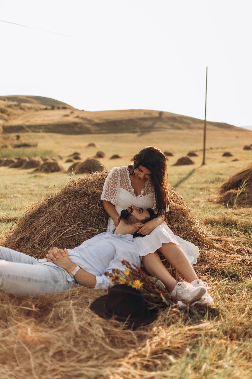 Affectionate pair cuddling on hay bale in sunlit meadow with rolling hills backdrop.