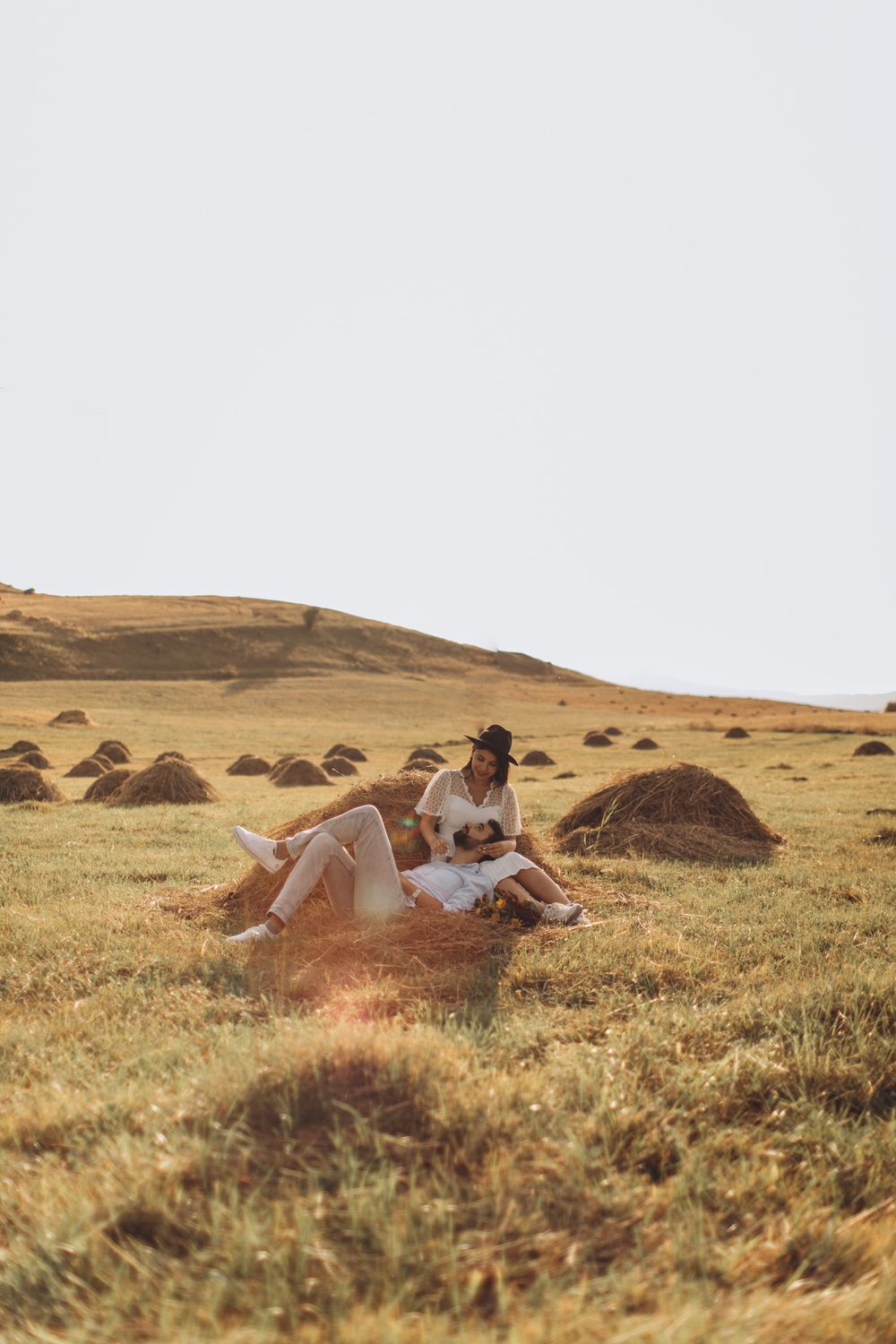 Candid couple's portrait in sun-drenched field with scattered hay bales.