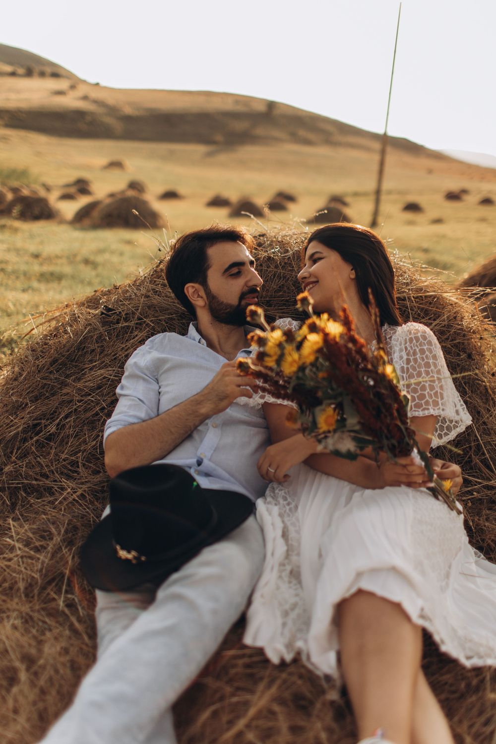 A romantic couple lounges on a hay bale in a golden field holding autumn flowers at sunset.