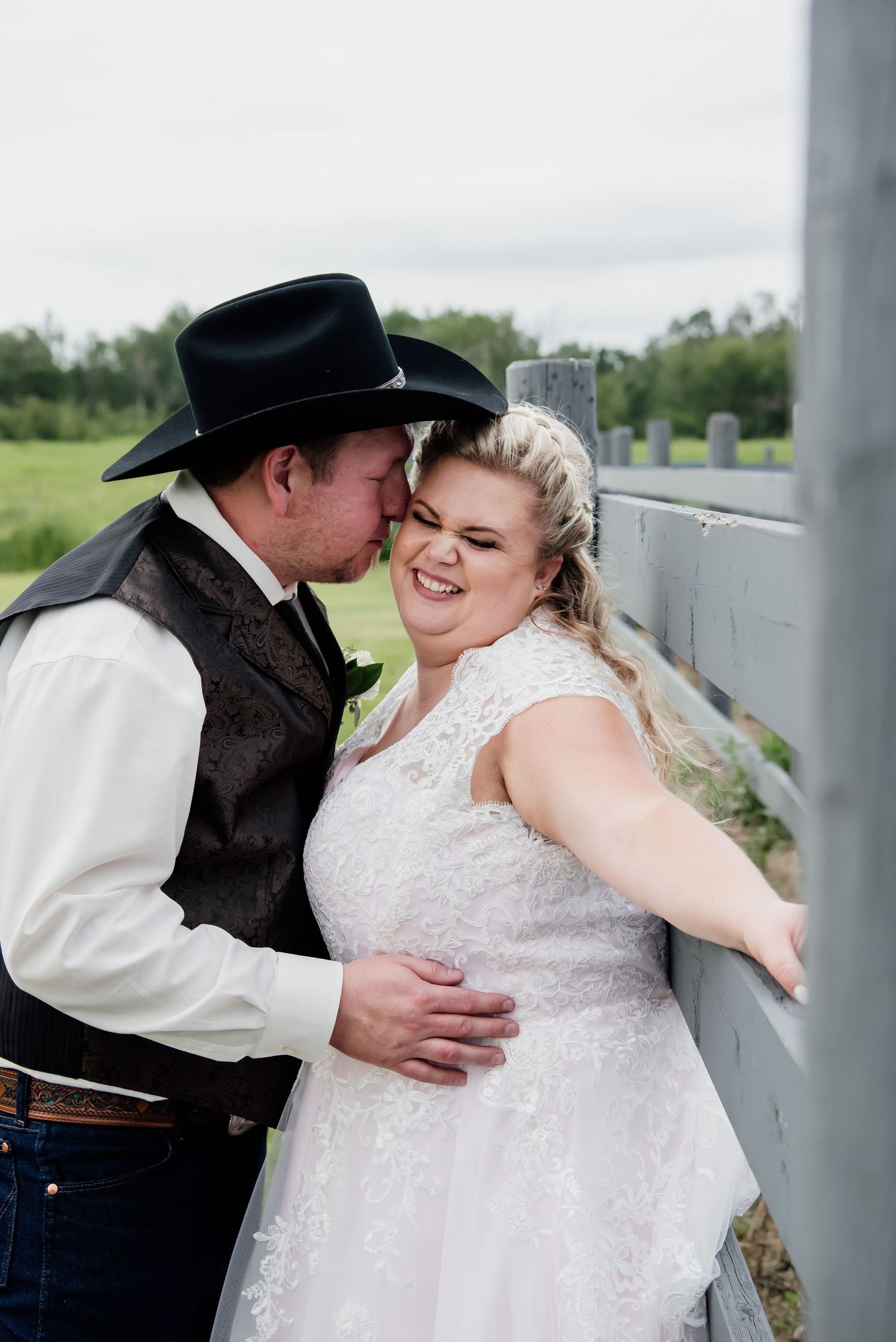 A couple shares a tender wedding moment against a white fence in western country attire.