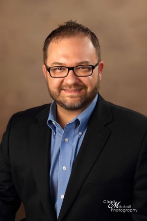 Professional business headshot of person in dark suit jacket, blue button-down shirt and glasses against brown backdrop.