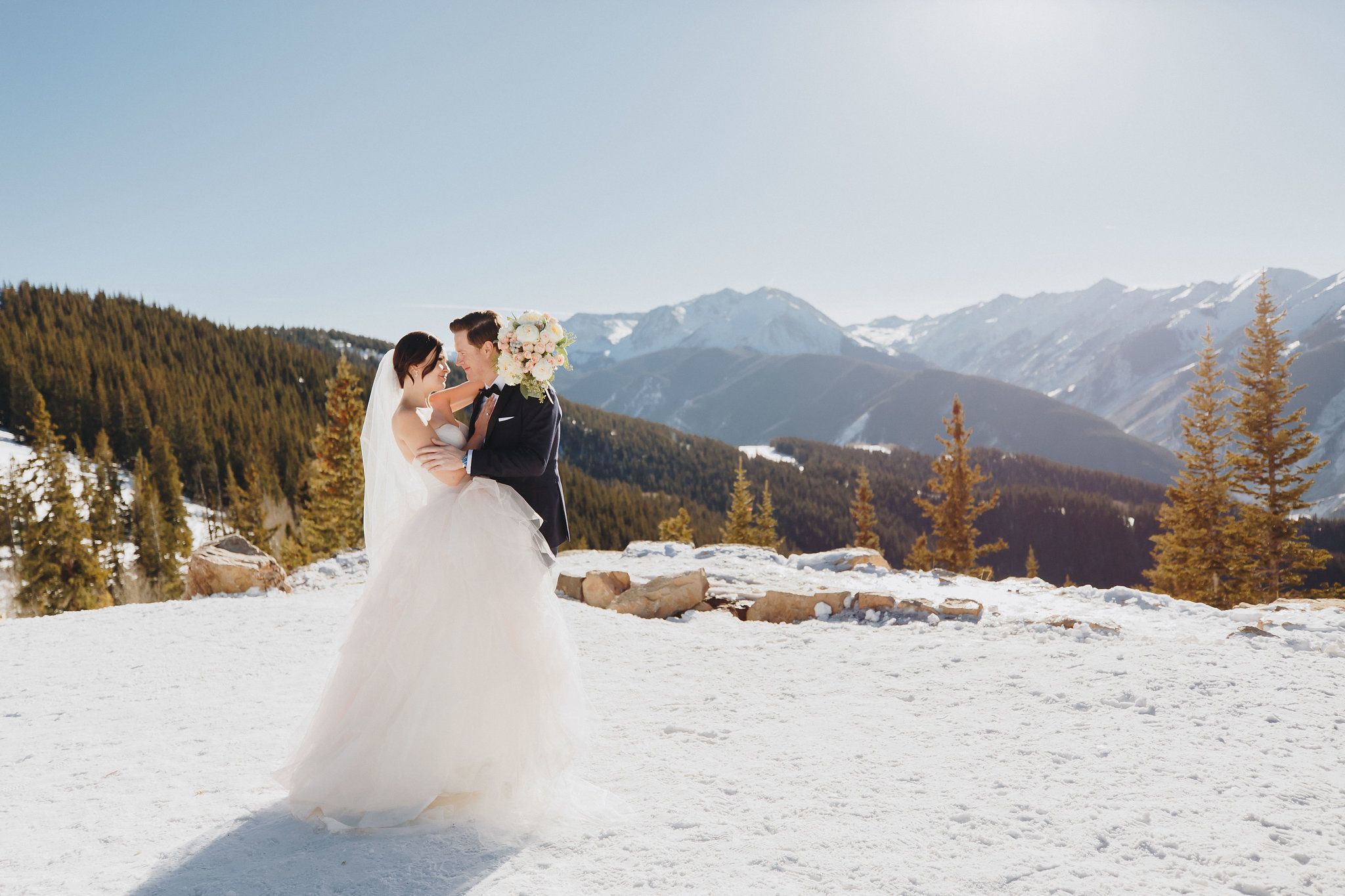 A romantic winter wedding portrait with snow-covered mountains and evergreen forest in the background.