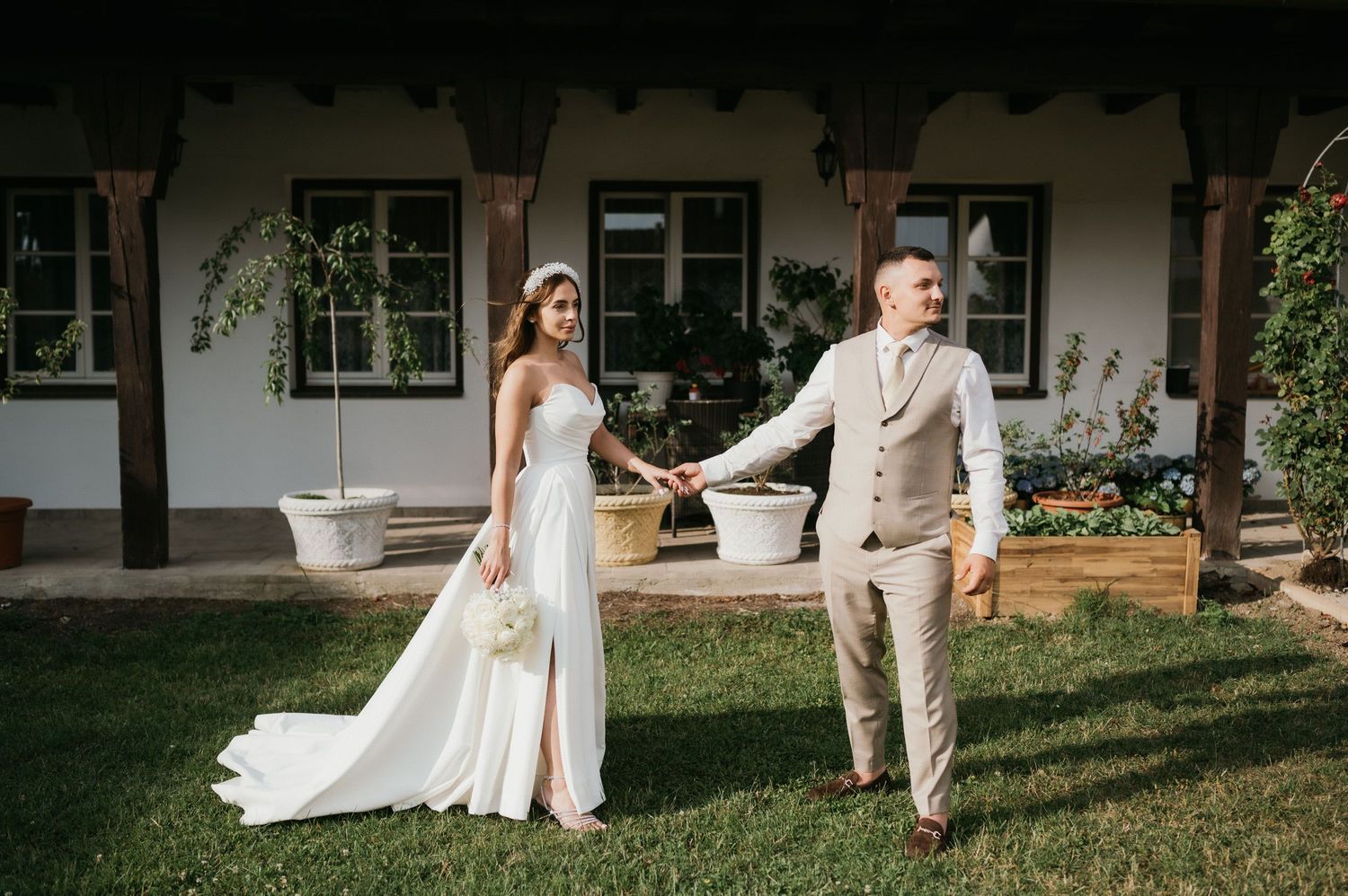 Couple in wedding attire walking hand in hand through a garden courtyard.