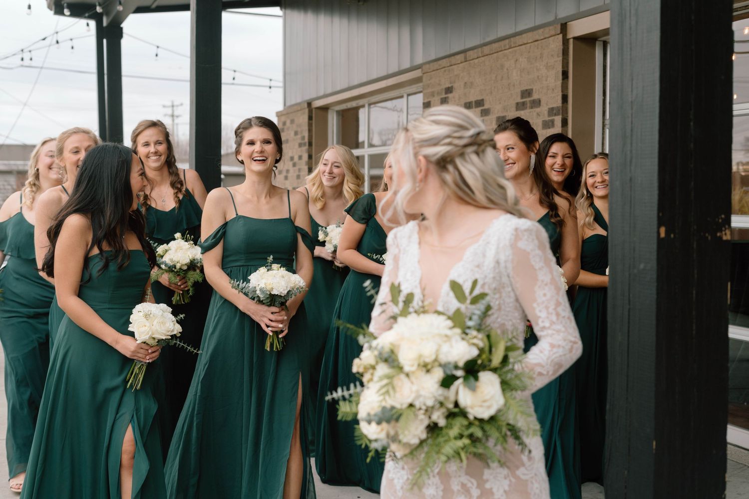 a bride in a white lace dress looks back at her bridesmaids in green dresses holding white boquets