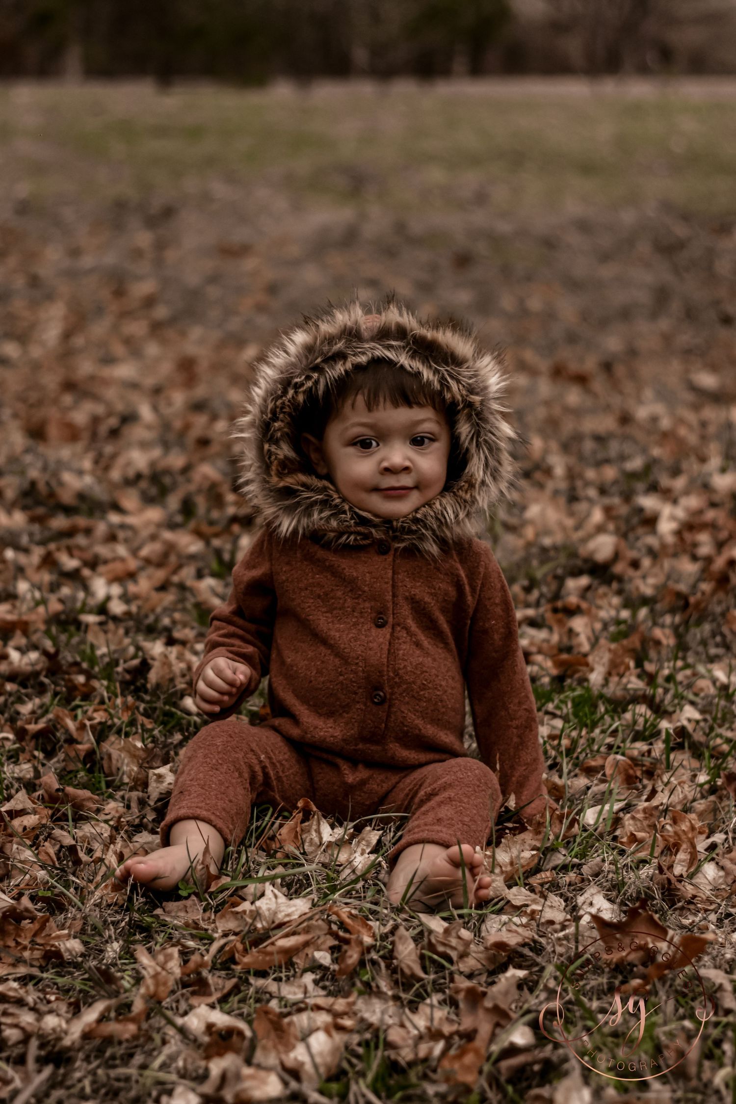 Baby wearing brown fuzzy bear outfit playing in fallen autumn leaves.
