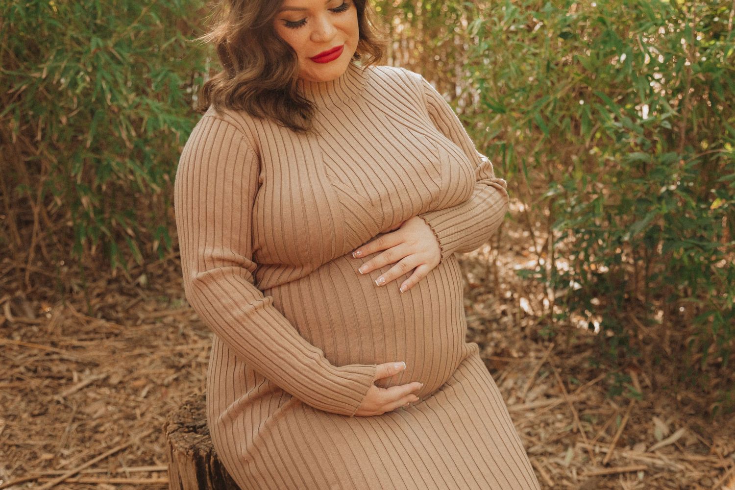 Maternity portrait featuring person in ribbed beige sweater dress against natural foliage.