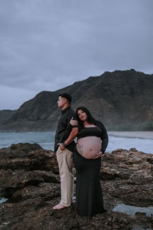 Expectant couple poses on rocky shore with dramatic mountain landscape behind them.