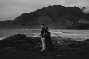 Silhouette of couple standing on rocky coastline with mountains in background at dusk.
