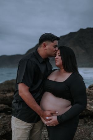 Maternity photo of couple embracing on rocky beach with mountains and ocean in background.